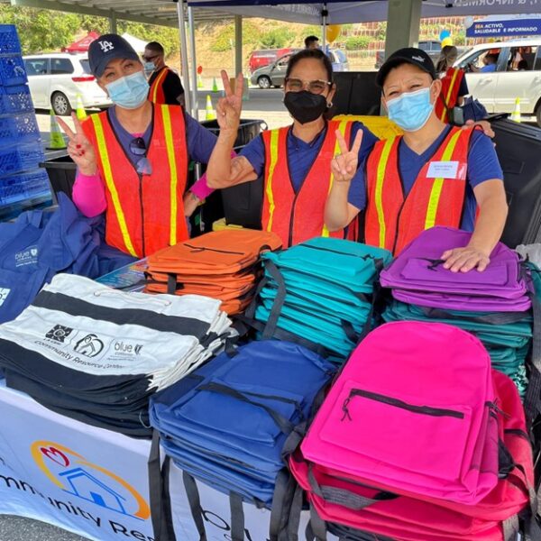 community resource center staff holding peace sign behind booth with backpacks